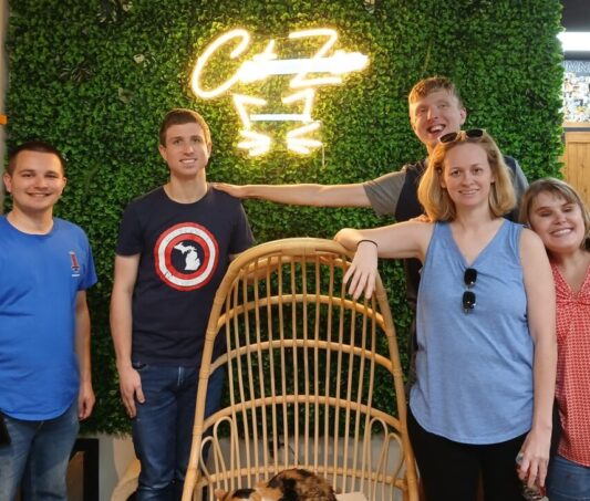 five participants stand around a cat sitting in a hanging chair, with a light up sign saying "cat Zen" behind it. Everyone is smiling with their arms around each other.