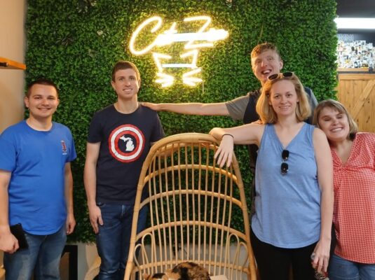 five participants stand around a cat sitting in a hanging chair, with a light up sign saying "cat Zen" behind it. Everyone is smiling with their arms around each other.